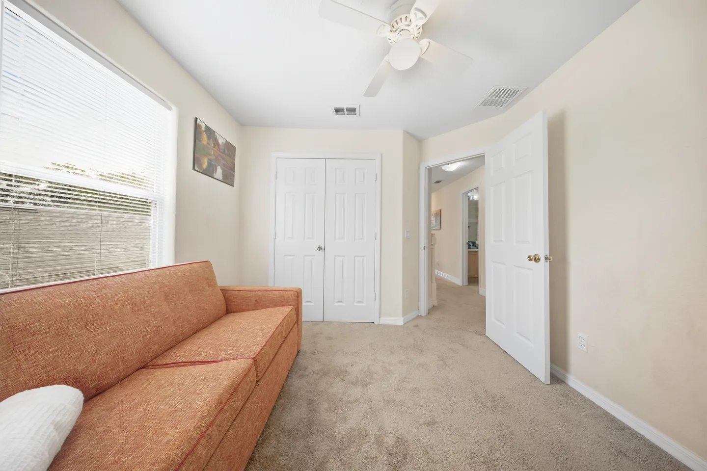 Carpeted room with an orange couch, white closet doors, a ceiling fan, and an open doorway showing a hallway.
