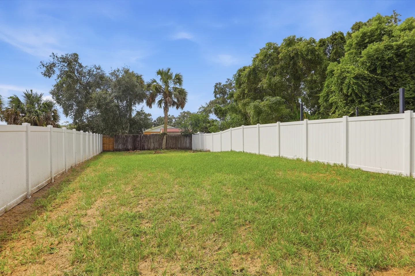 A large, grassy backyard enclosed by white vinyl privacy fences with trees and a blue sky in the background.