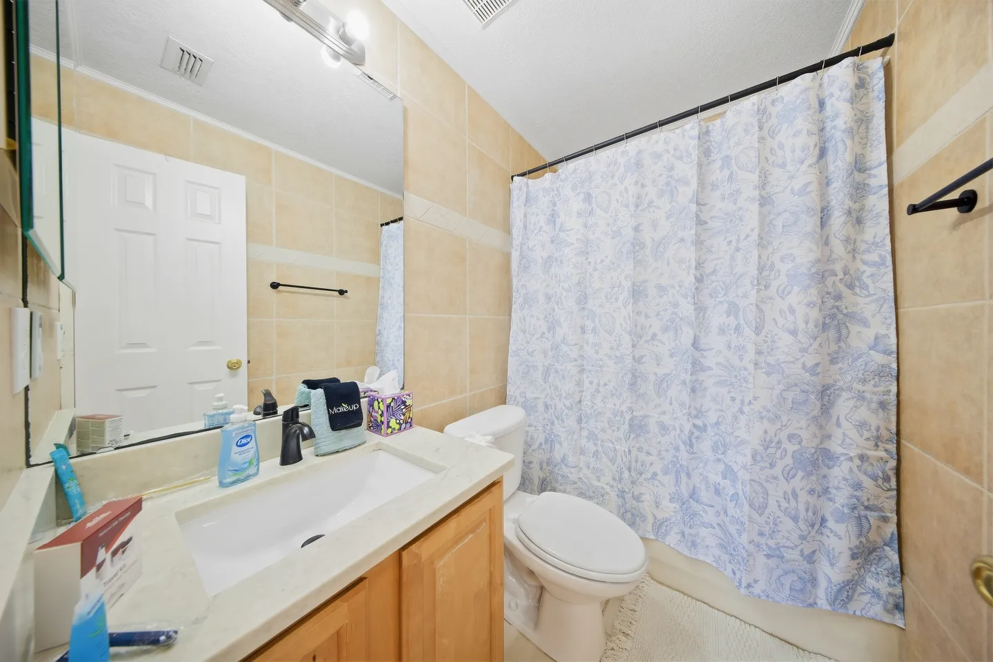 Bathroom with light wood vanity, beige tiled walls, white toilet, and a blue and white patterned shower curtain.