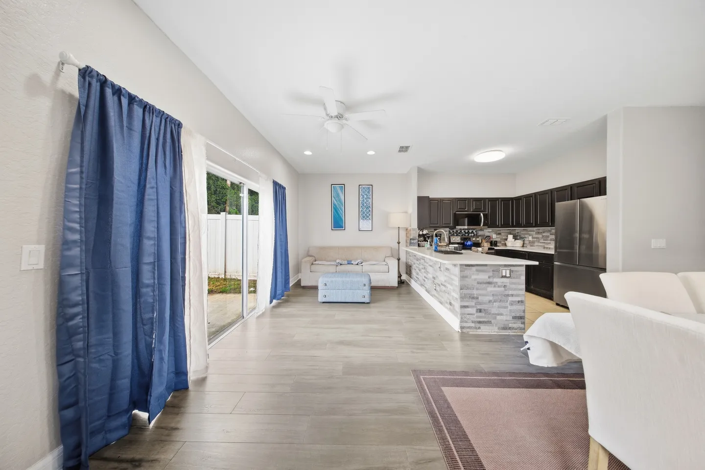 Open-concept living room and kitchen with wood-look flooring, dark cabinetry, and sliding glass doors with blue curtains.