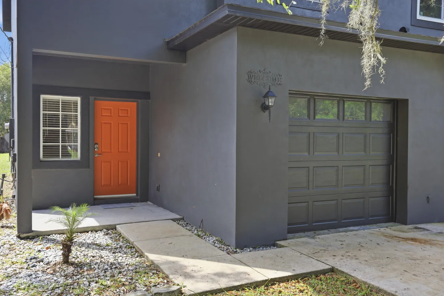 Exterior front entrance of a charcoal gray house featuring an orange front door, a window with blinds, and an adjacent garage door.