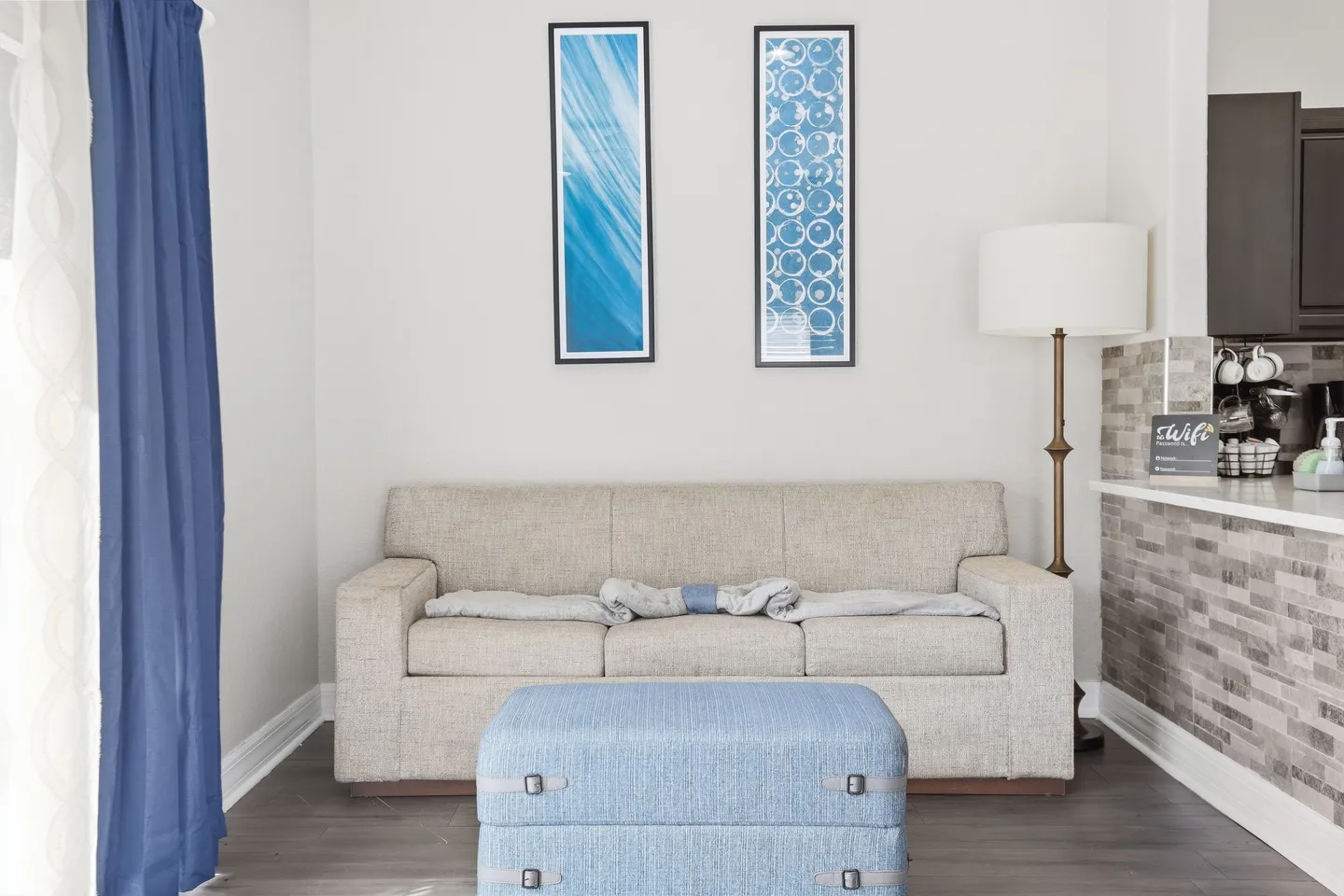 Living area with a neutral fabric sofa, light blue ottoman, floor lamp, and framed blue wall art.