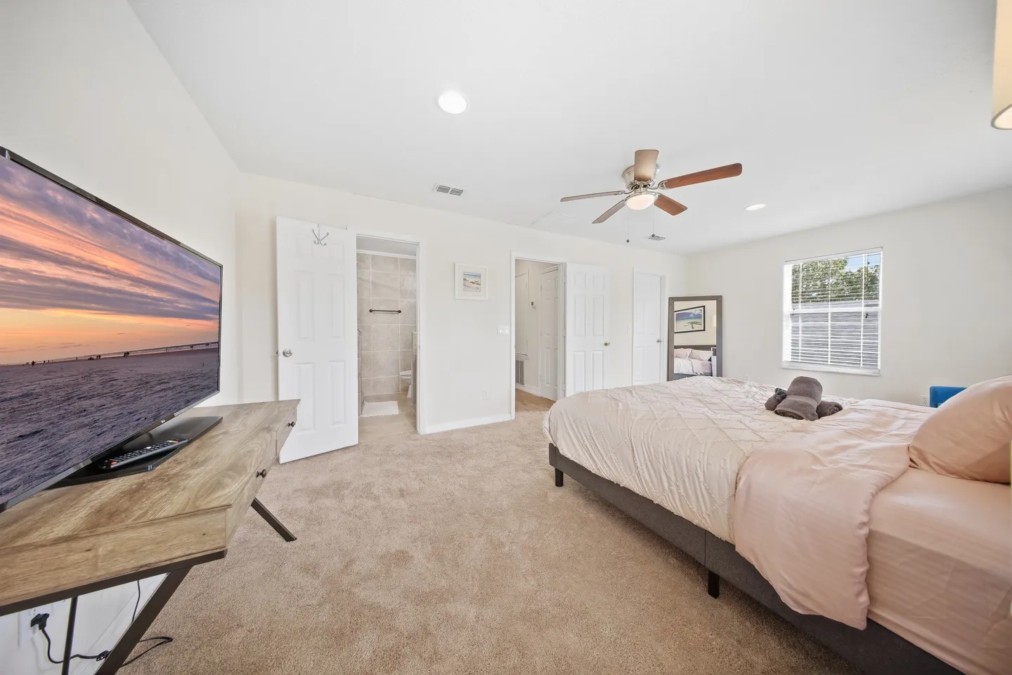 Bedroom featuring carpeted flooring, a made bed, a ceiling fan, a wall-mounted flat-screen TV on a wooden console, and doorways leading to a bathroom and hallway.