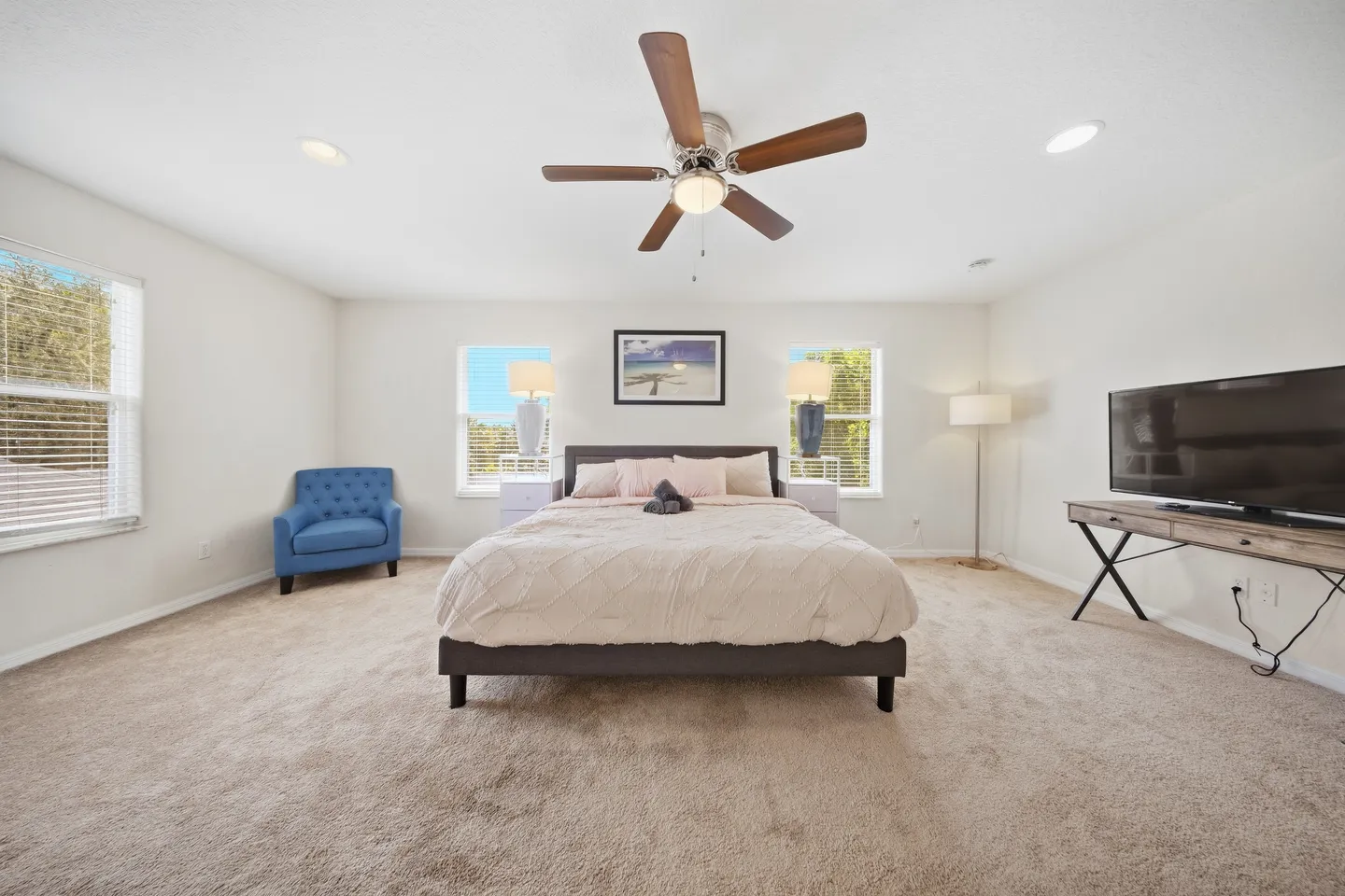 Spacious bedroom with carpeted floor, large bed, ceiling fan, armchair, and television on a console table.