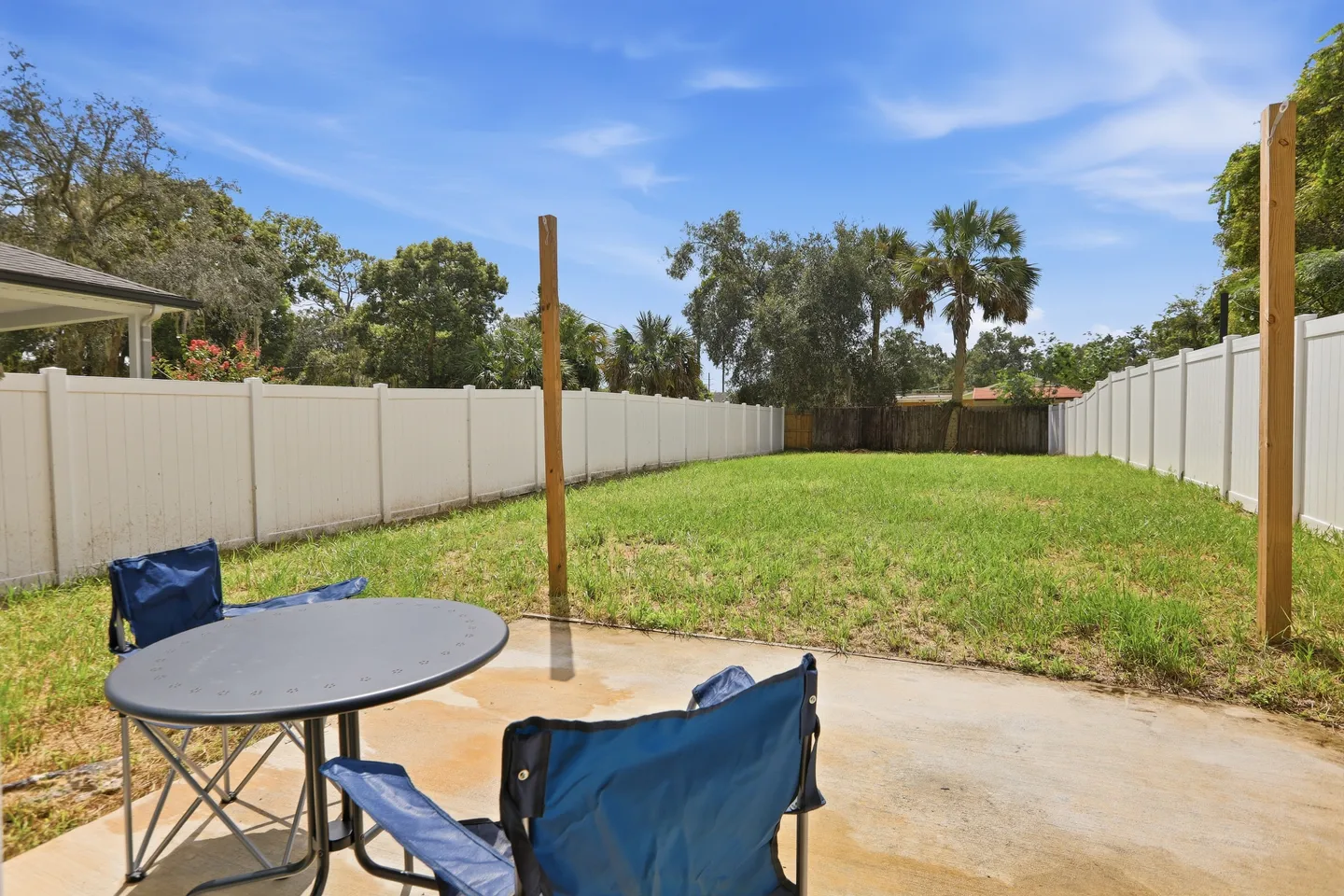 Fenced grassy backyard with a concrete patio featuring folding chairs and a small round table.