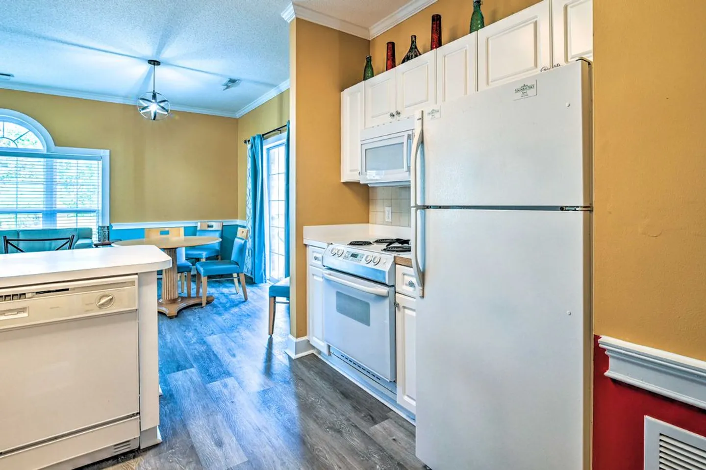 Kitchen with white appliances and cabinets, leading into a dining area with a round table and turquoise chairs.