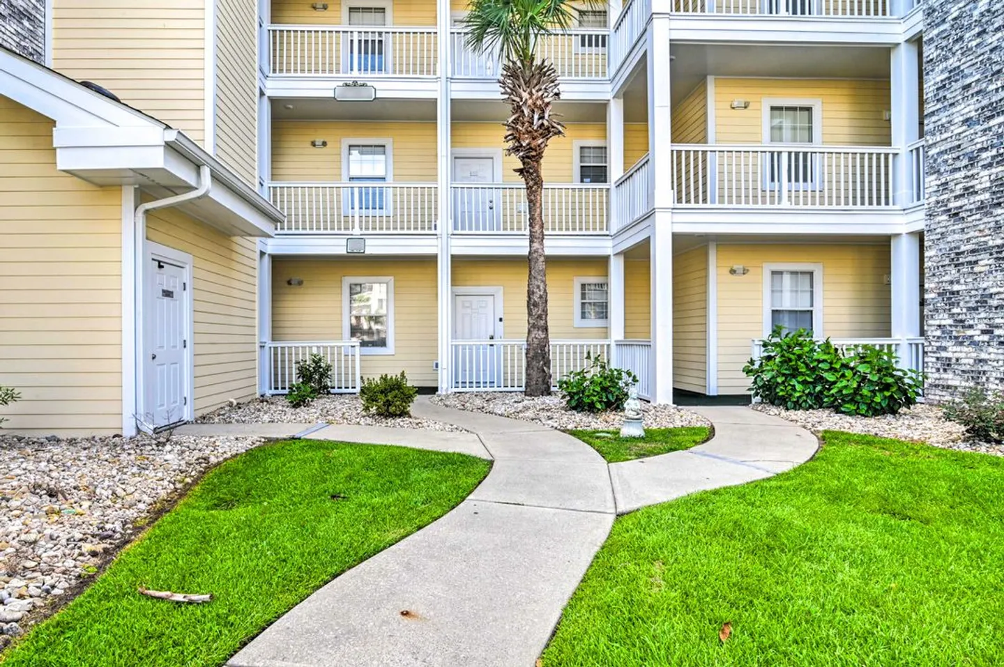 Exterior view of a multi-story apartment building with yellow siding, white railings, a concrete walkway, green lawn, and a palm tree.