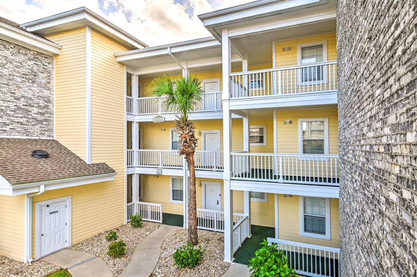 Exterior of a three-story apartment or condominium building with yellow siding, white railings, and a small palm tree in the courtyard.