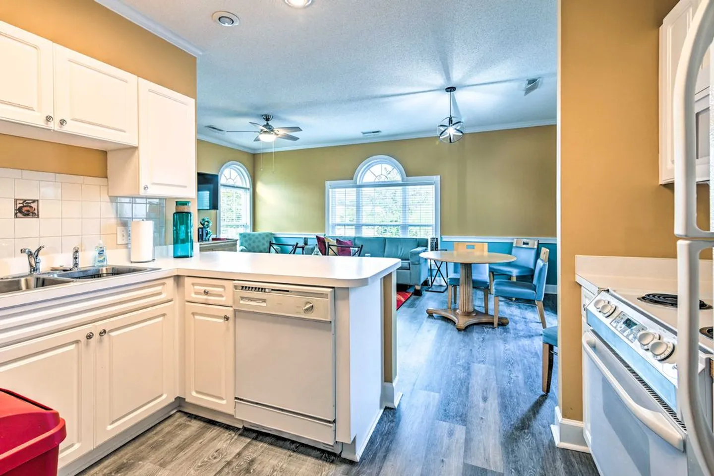 View from a kitchen with white cabinets looking into an open dining and living area with wood-look flooring and arched windows.