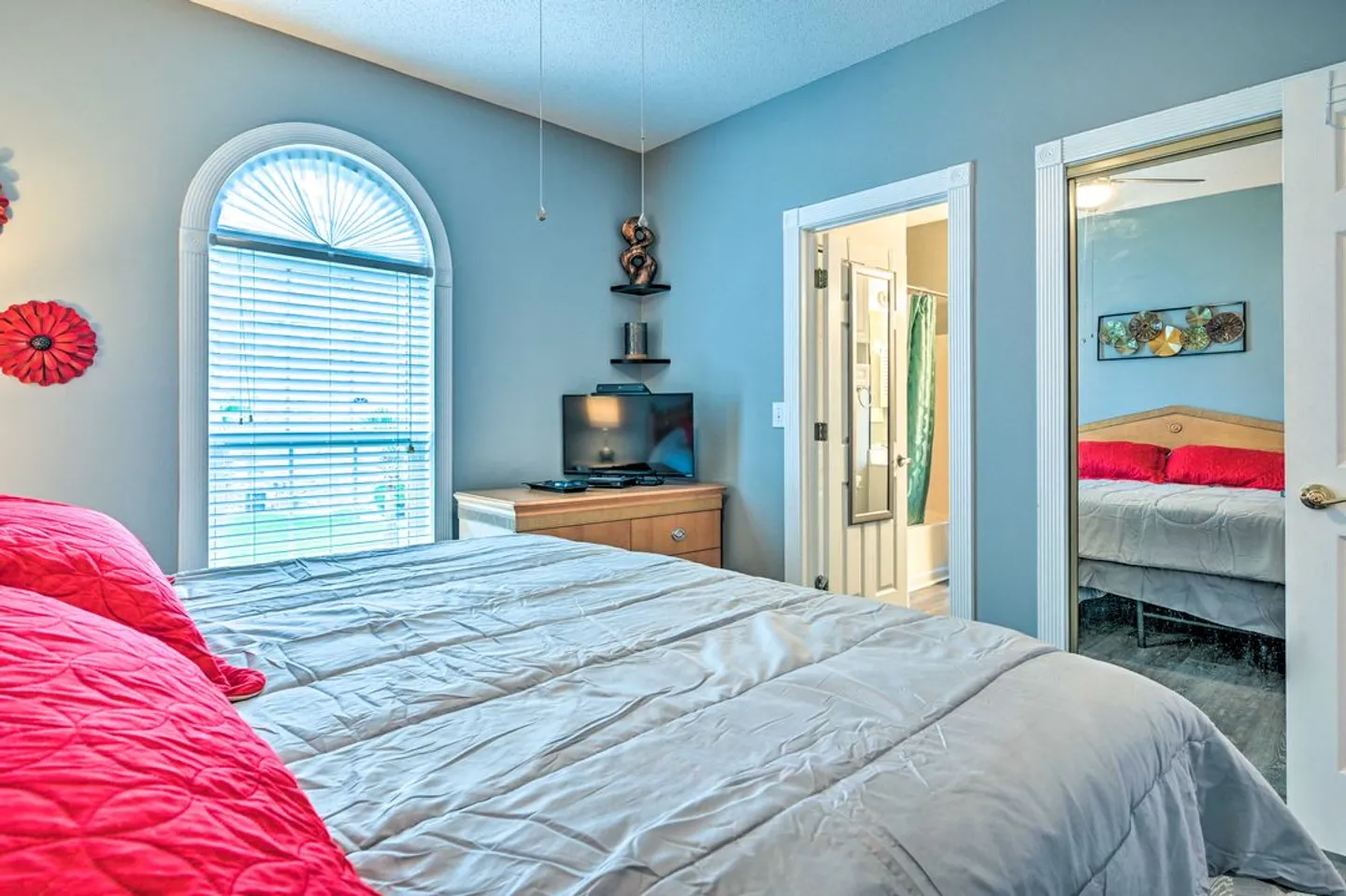 Bedroom with blue walls, an arched window with blinds, a dresser with a television, and an ensuite bathroom door.
