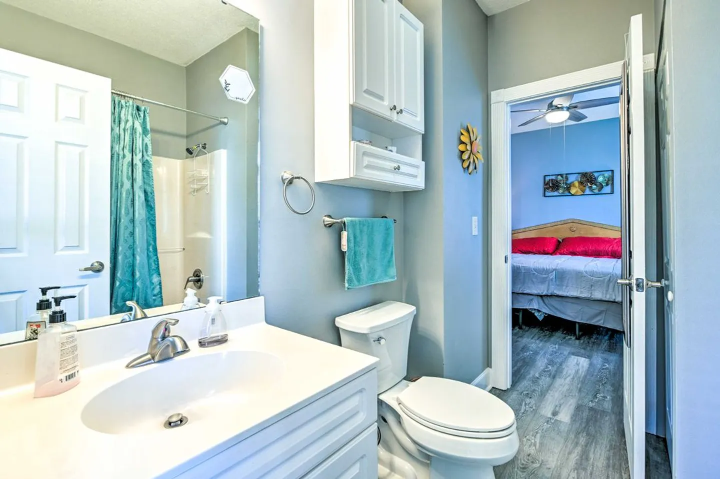 Bathroom with white vanity, mirror, toilet, overhead storage cabinet, and doorway leading to a bedroom.