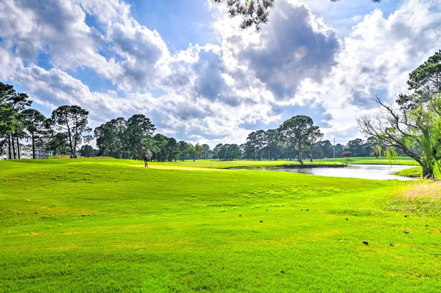 A golf course with a green lawn, mature trees, a water hazard, and a partly cloudy blue sky.