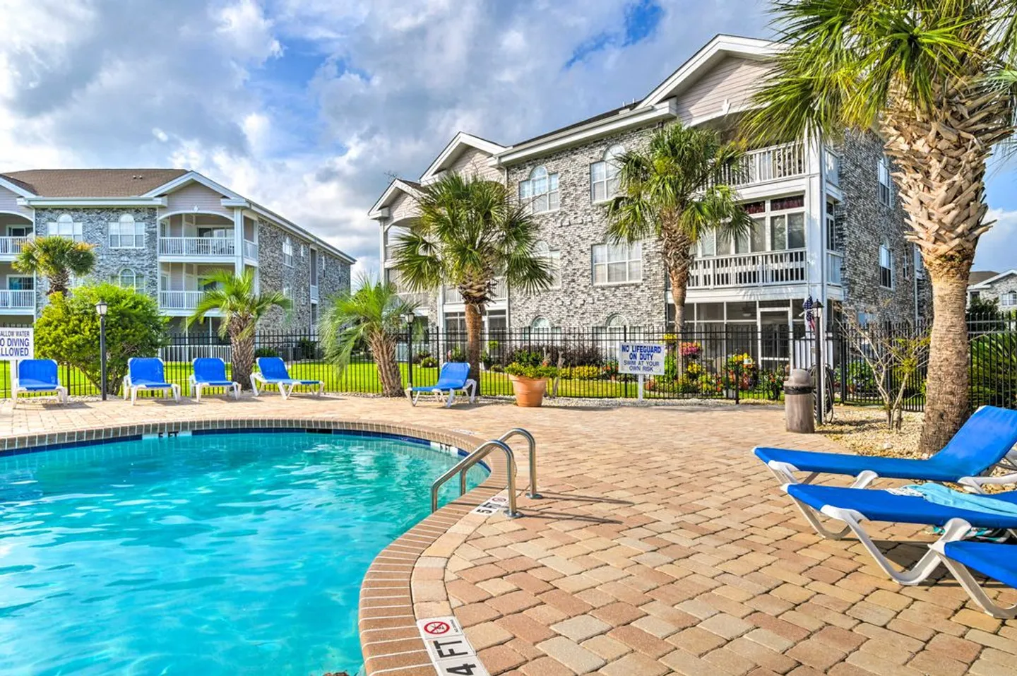 Outdoor swimming pool surrounded by a brick paver patio and lounge chairs near multi-story residential buildings.