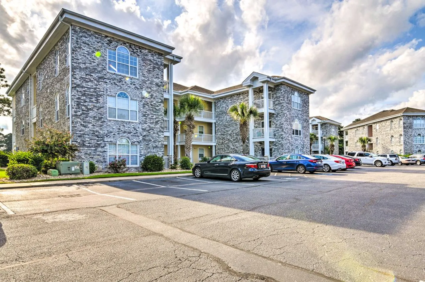 Exterior view of a multi-story stone facade apartment building with balconies and a paved parking lot with cars in front.