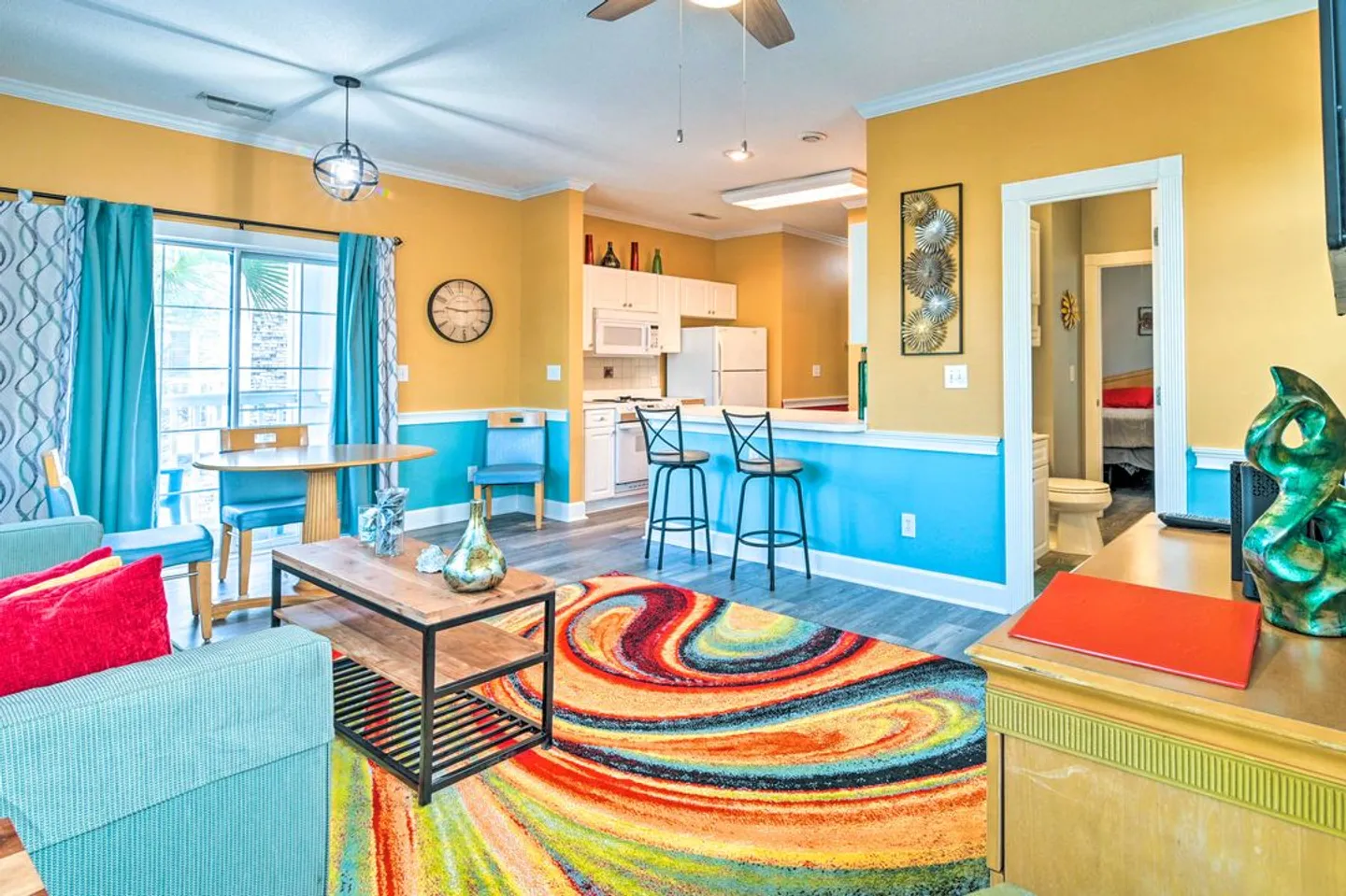 Open living area with yellow and bright blue walls, a vibrant swirling area rug, a coffee table, and a view into the kitchen with a breakfast bar.