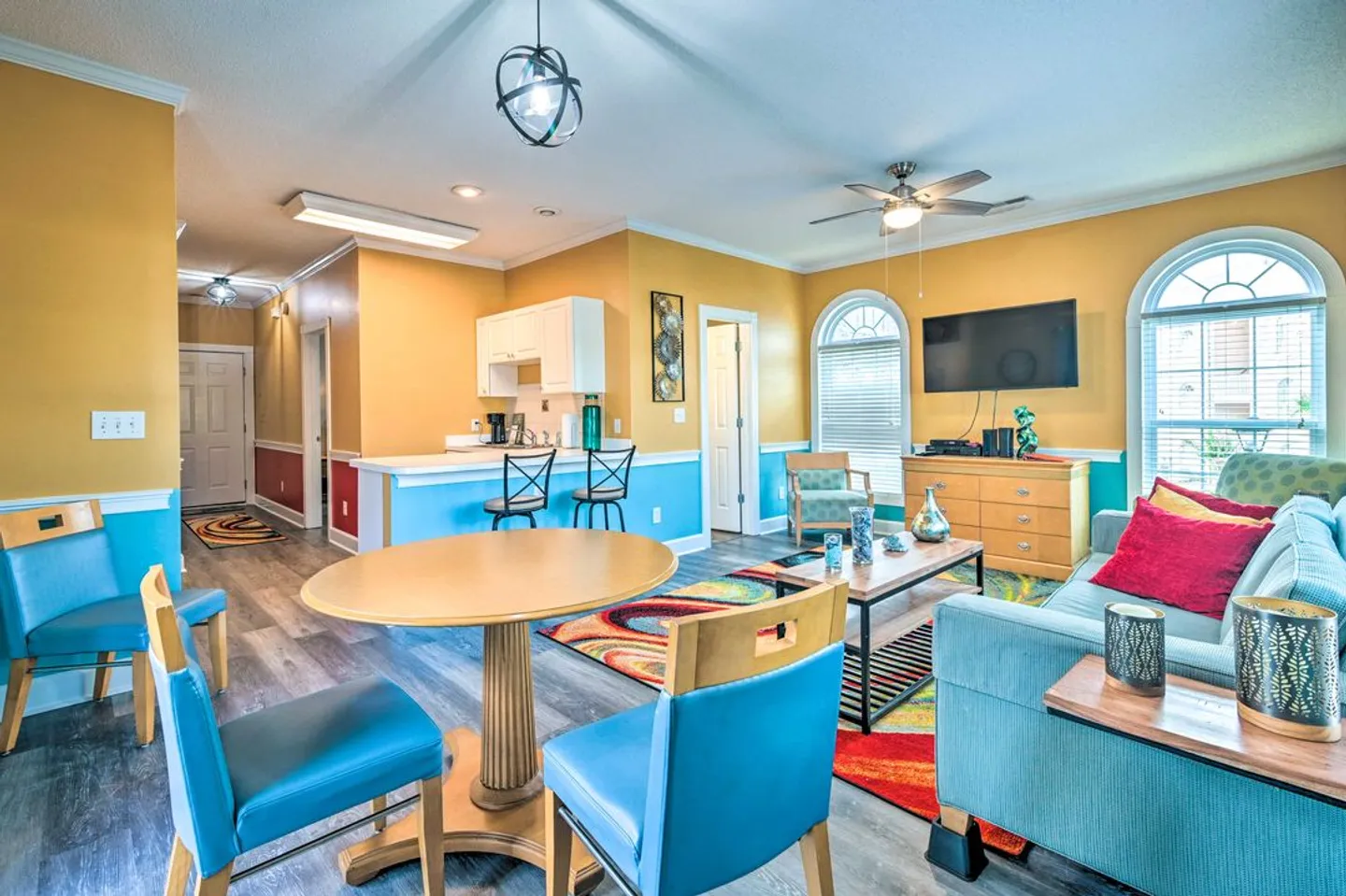 Open-concept living room and dining area featuring vibrant yellow and turquoise walls, wood-look flooring, a round table with blue chairs, and a seating area with a sofa and television.