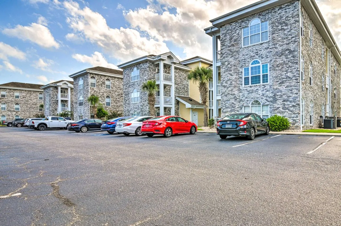 Exterior view of multi-story stone-faced apartment buildings bordering an open parking lot with parked cars under a blue sky.