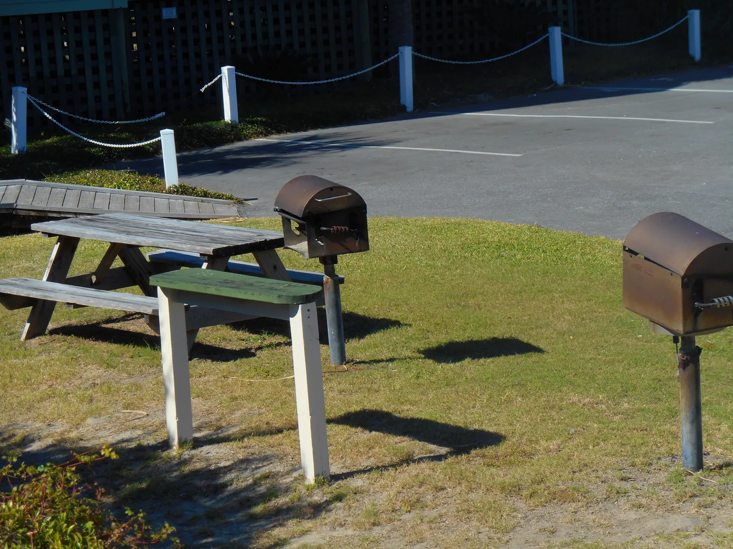 Outdoor picnic area with two park-style charcoal grills, a wooden picnic table, and a small prep table on a grassy patch beside a parking area.