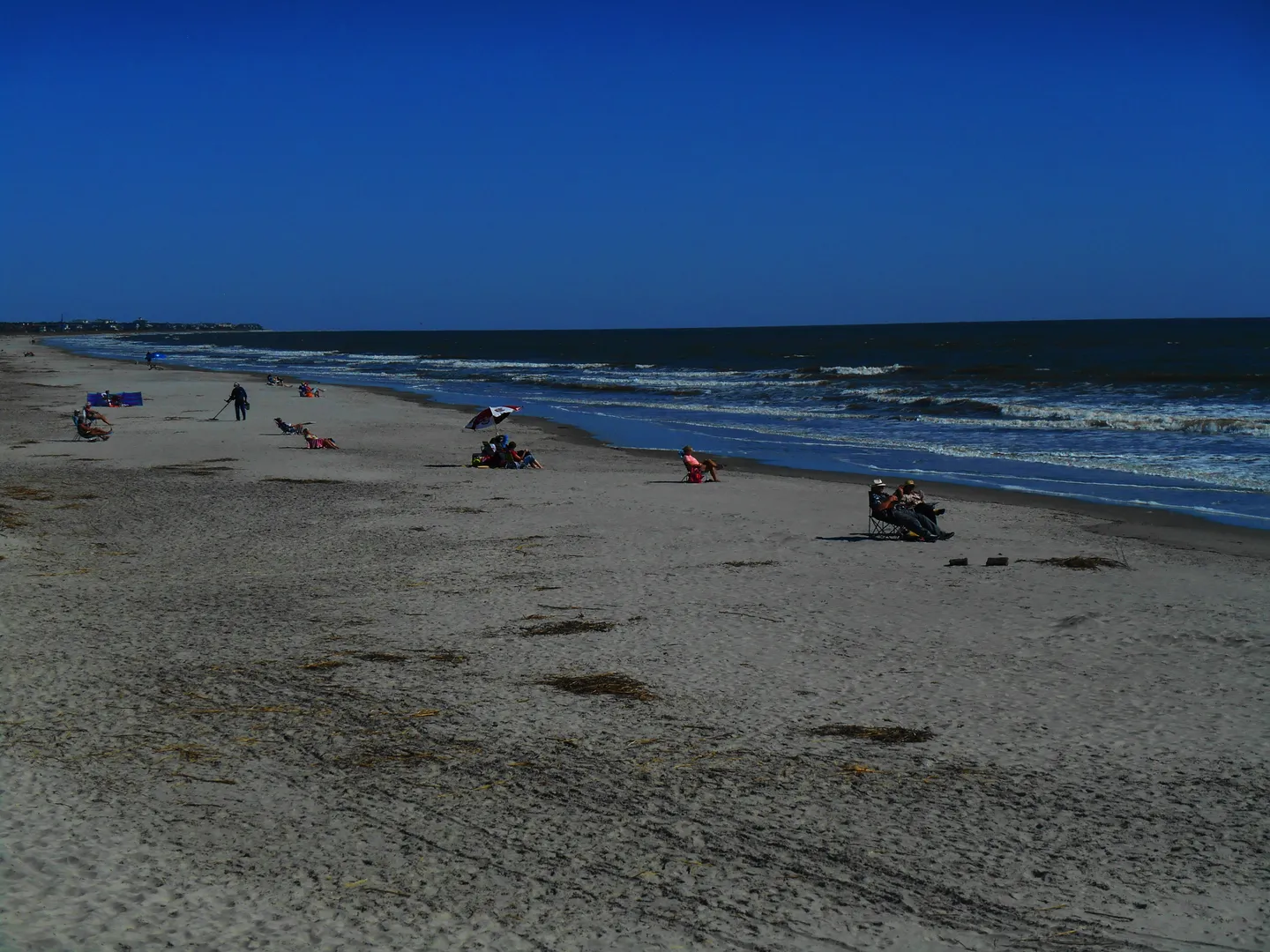 A sandy beach shoreline with people relaxing under a clear blue sky as ocean waves roll in.