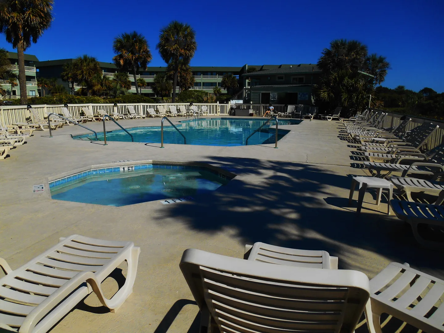 An outdoor community swimming pool and hot tub surrounded by lounge chairs and palm trees with residential buildings in the background.