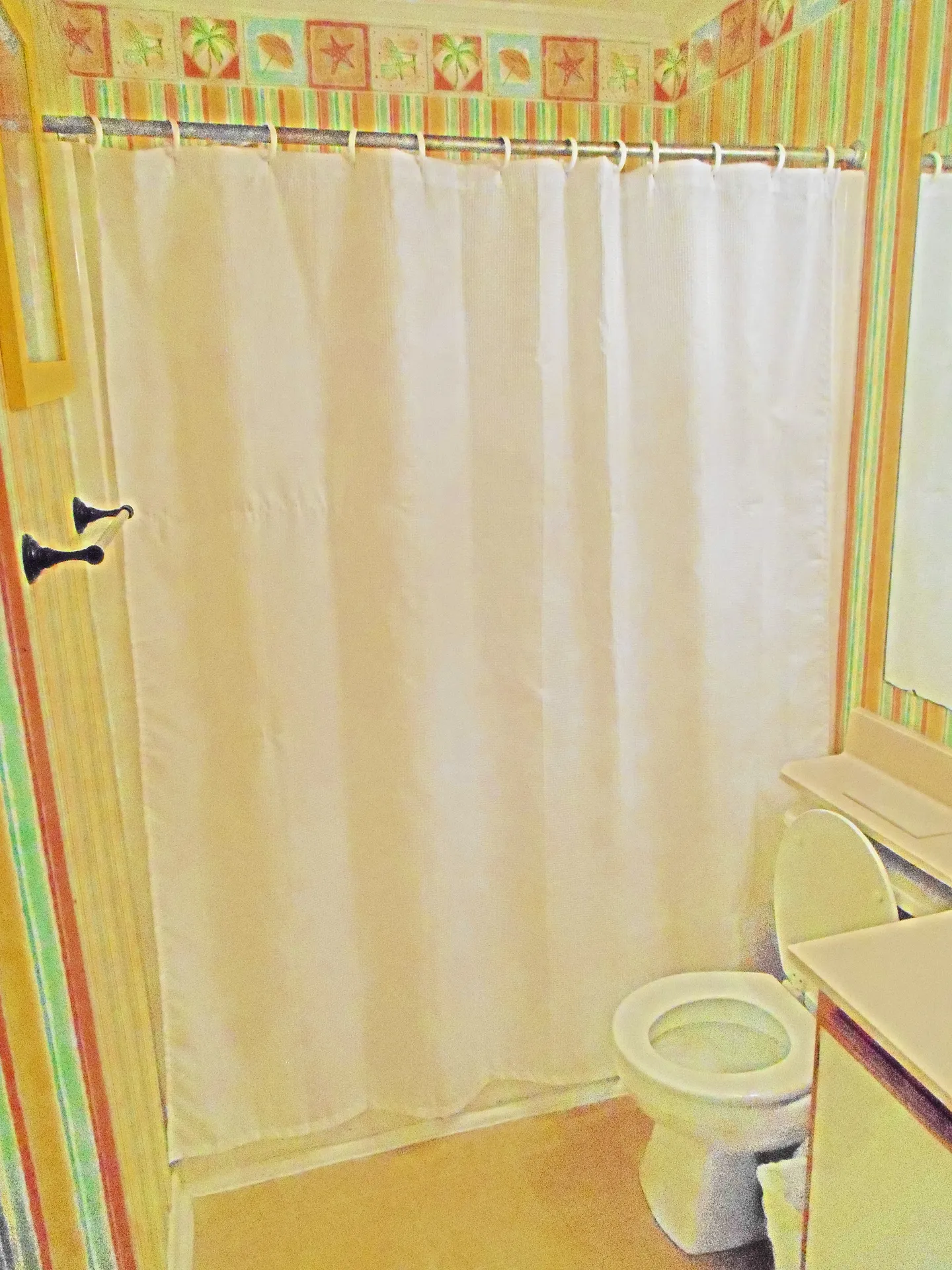 Bathroom showing a white shower curtain, a toilet, vanity corner, and striped wallpaper with a beach-themed border.