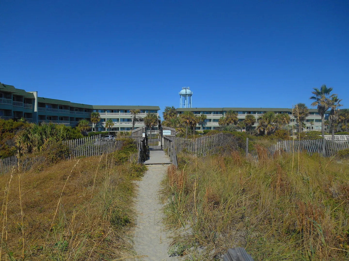 Sandy path leading through dunes and a wooden boardwalk toward a multi-story condominium complex under a clear blue sky.