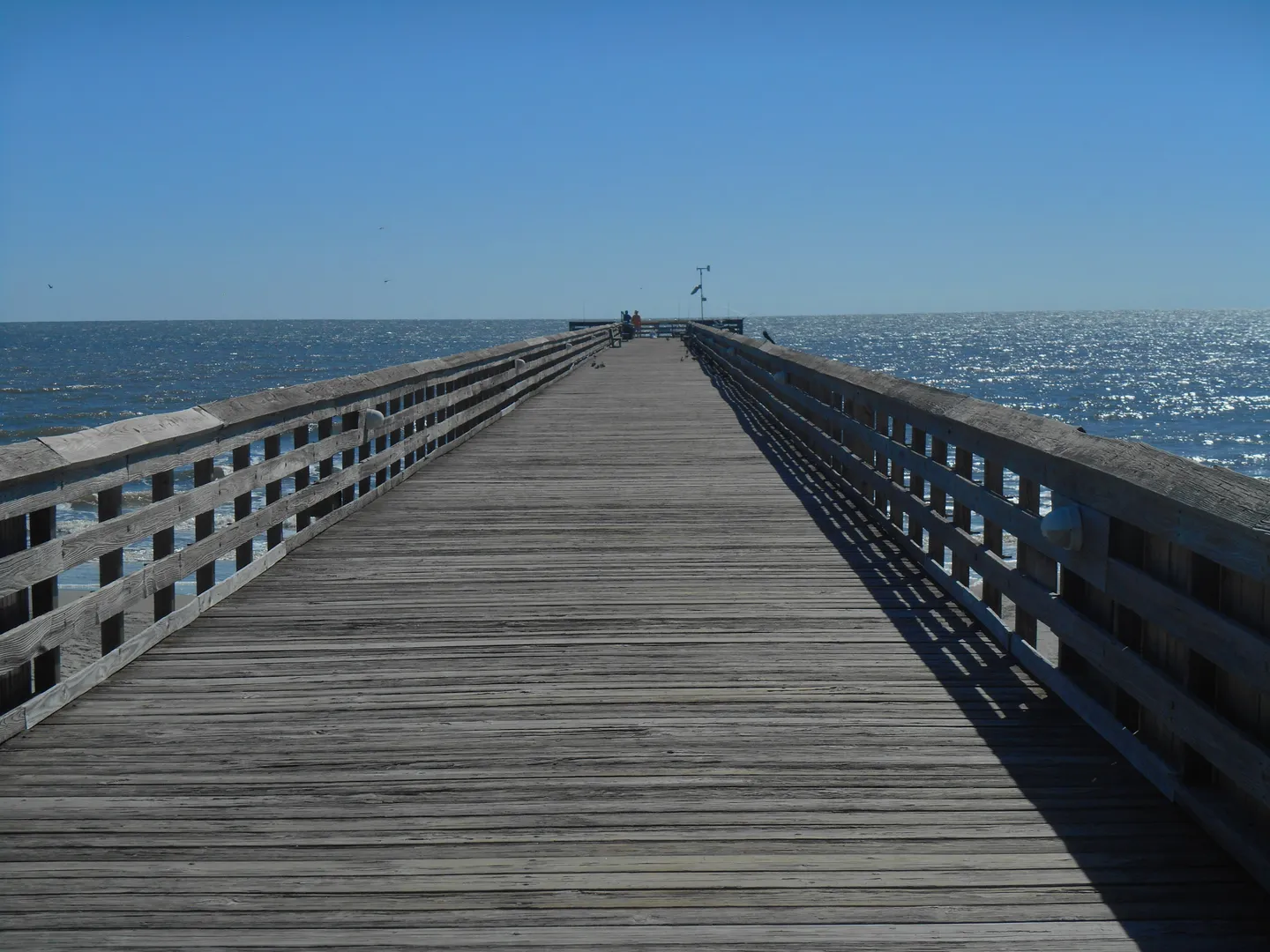 A long wooden pier extending over the ocean beneath a clear blue sky.