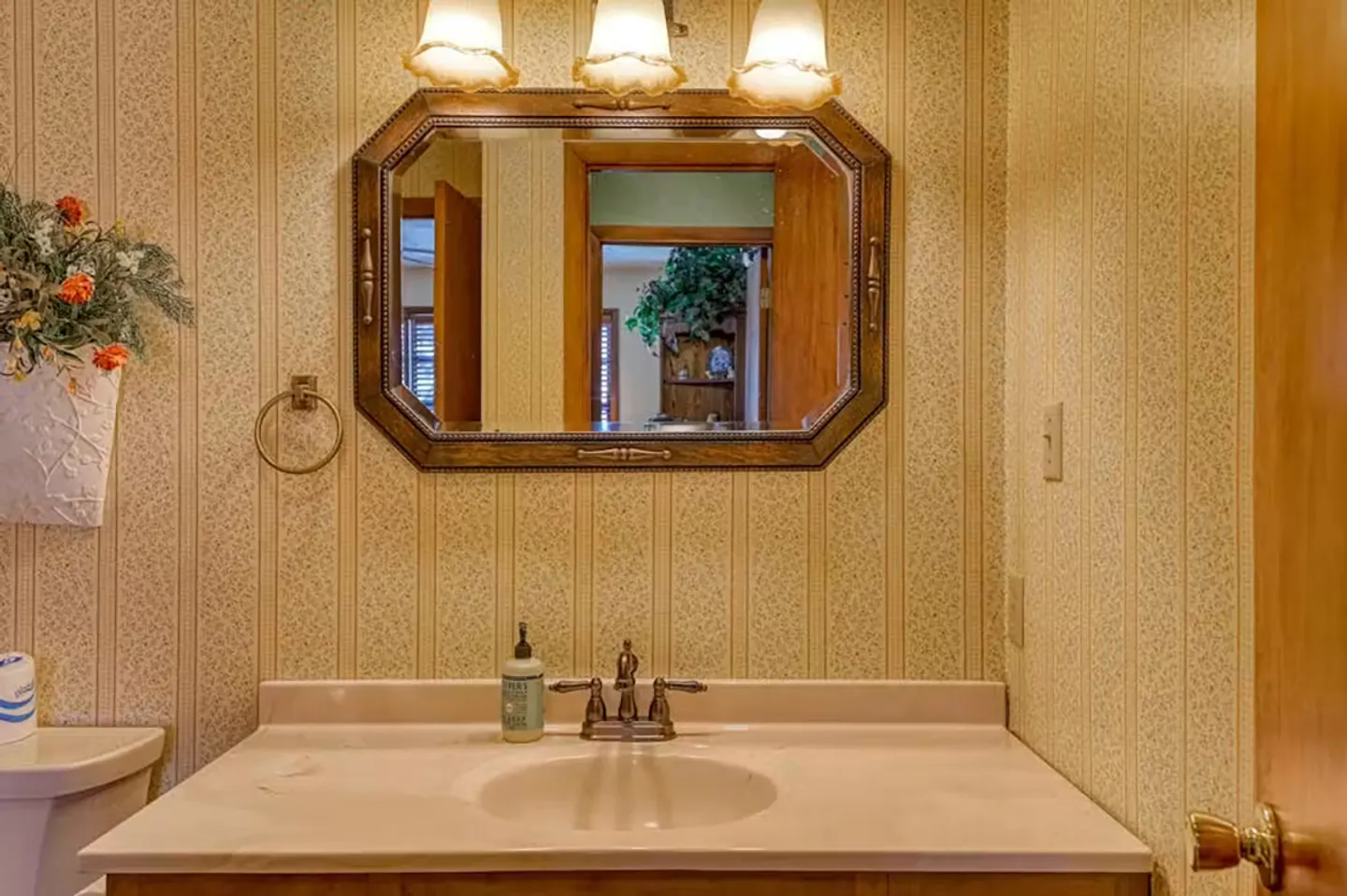 Bathroom vanity with sink, framed mirror, triple light fixture, and patterned wallpaper.