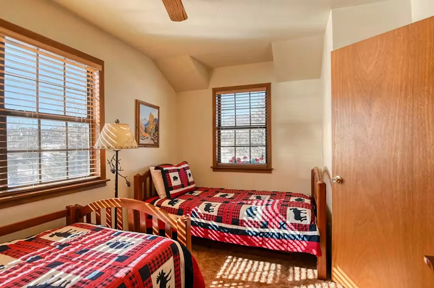 Bedroom with two wooden twin beds made with red plaid quilts and windows with blinds.