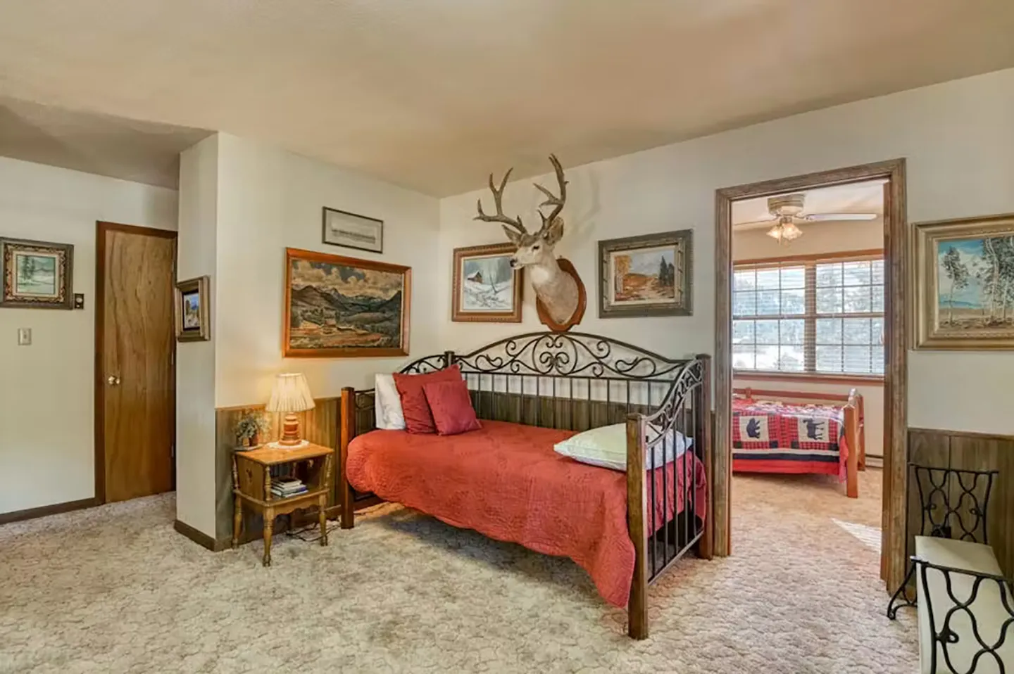 A carpeted bedroom featuring a rustic daybed, a mounted deer head, framed artwork, and a doorway leading into an adjoining bedroom.