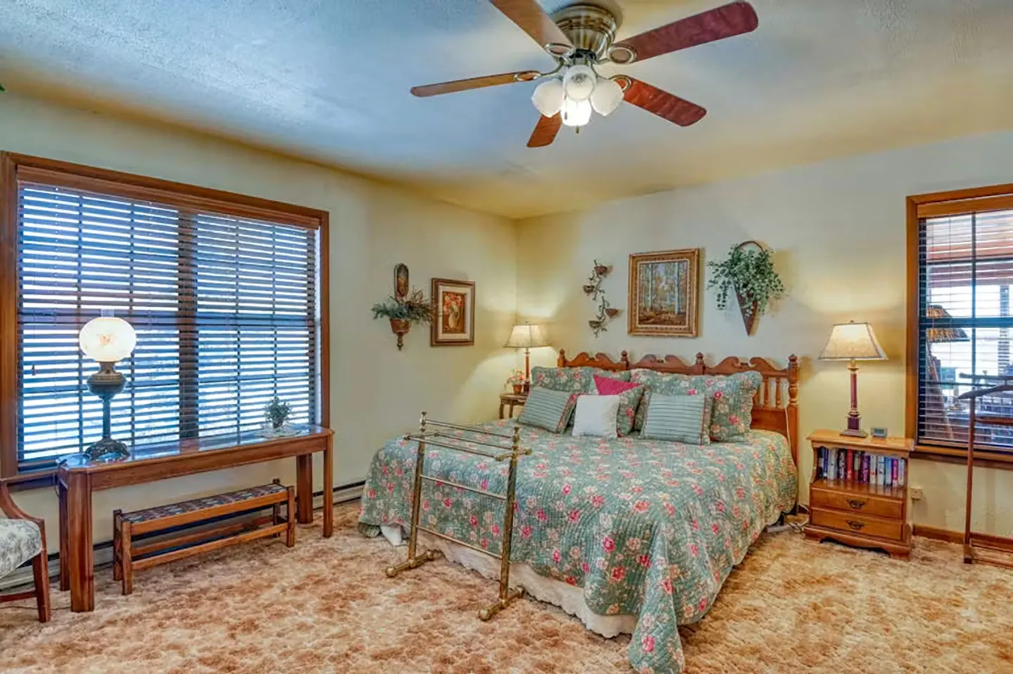 Bedroom with patterned brown carpet, a floral bedspread, wooden headboard, side tables with lamps, and windows with blinds.