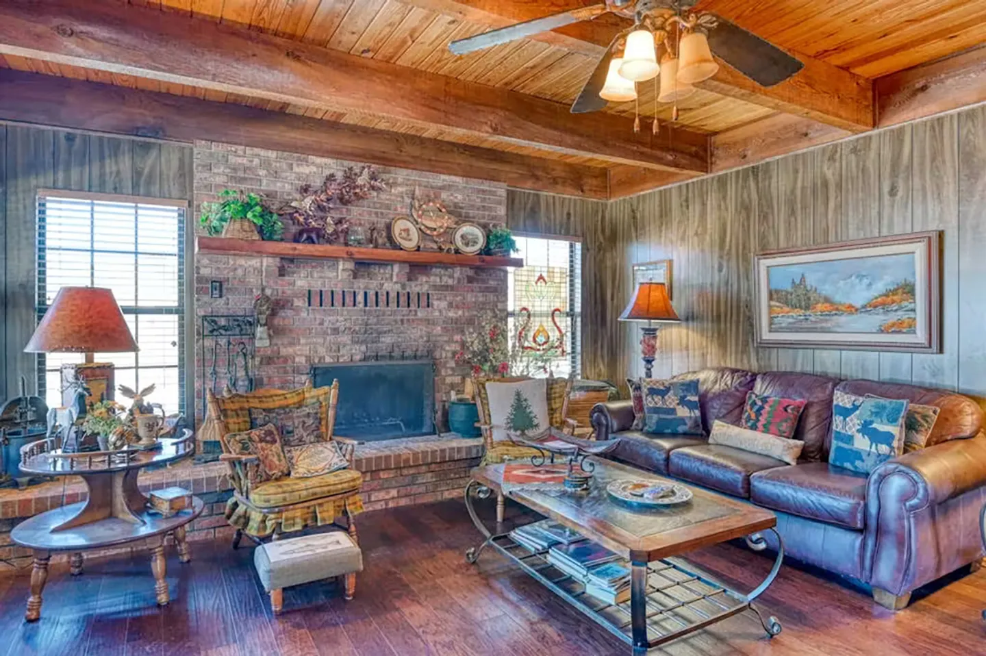 Rustic living room with a brick fireplace, exposed wood ceiling beams, leather sofa, and wood-paneled walls.