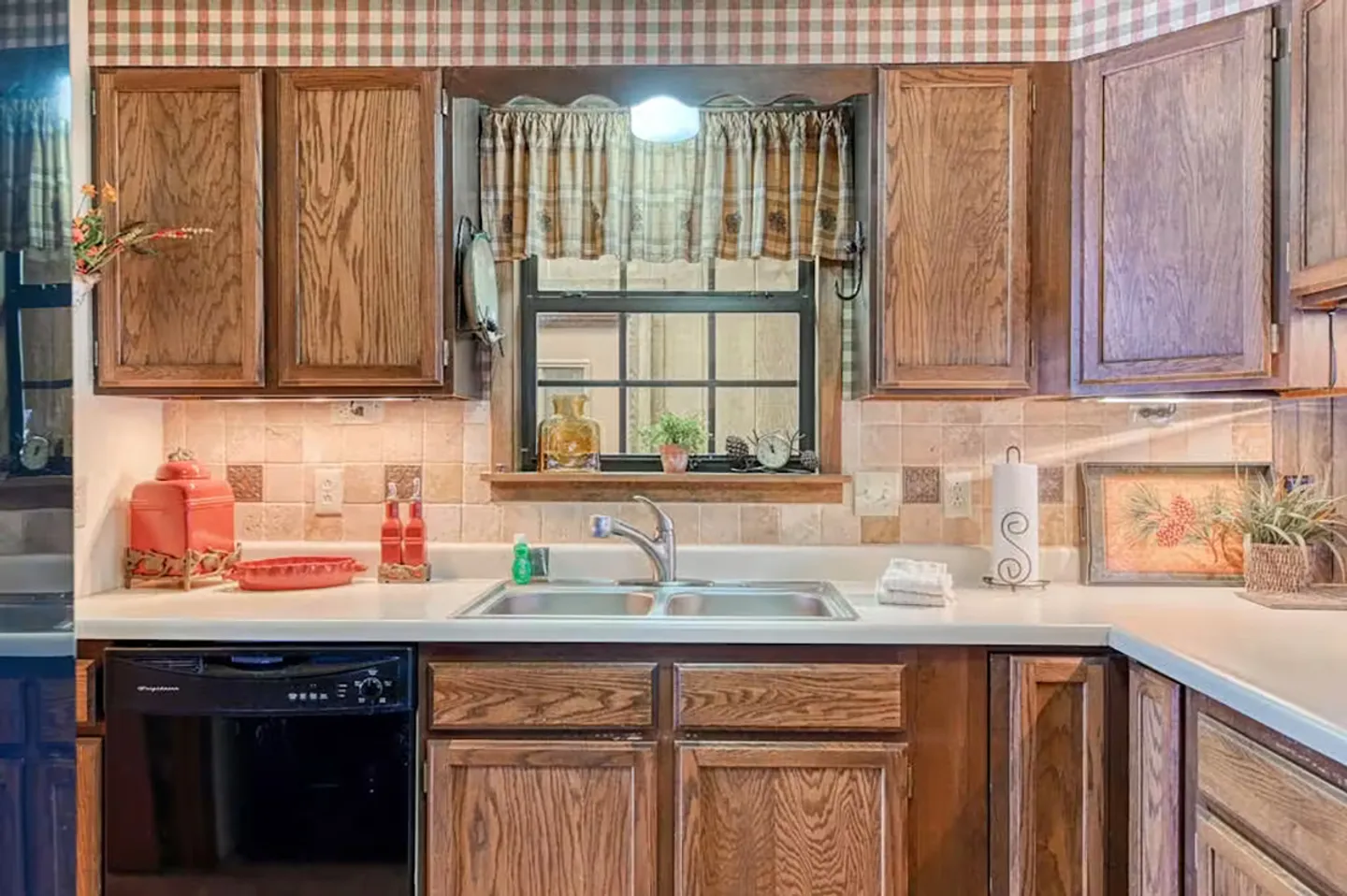 Close-up of a kitchen sink area featuring wooden cabinets, a double stainless steel sink, tiled backsplash, and plaid wallpaper border.