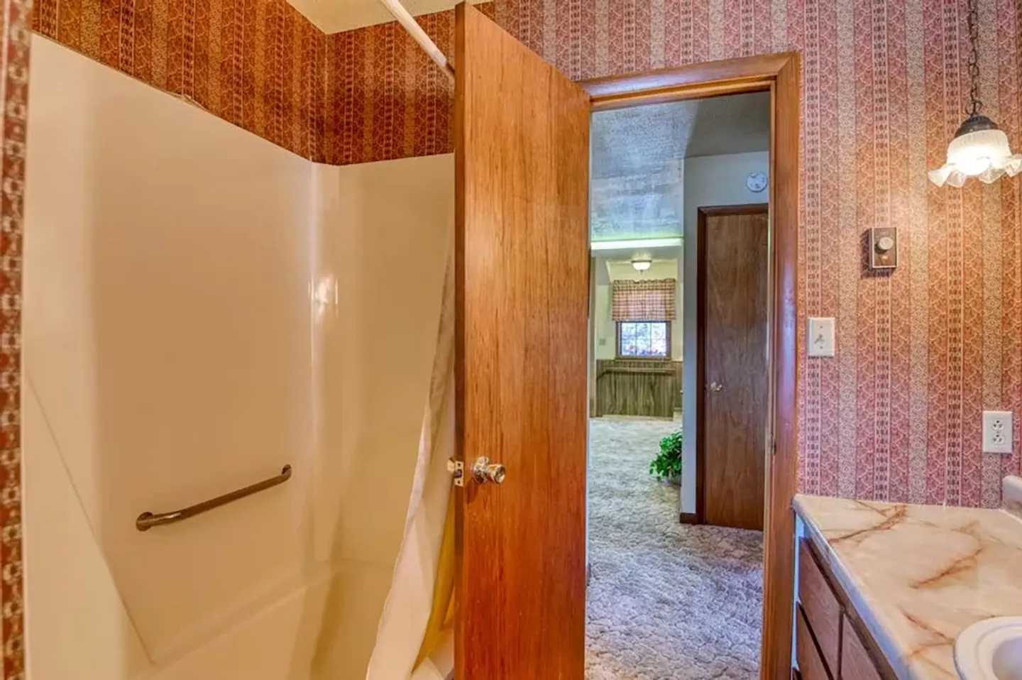Bathroom with patterned wallpaper, a tub and shower stall, and an open doorway looking out to a carpeted room.