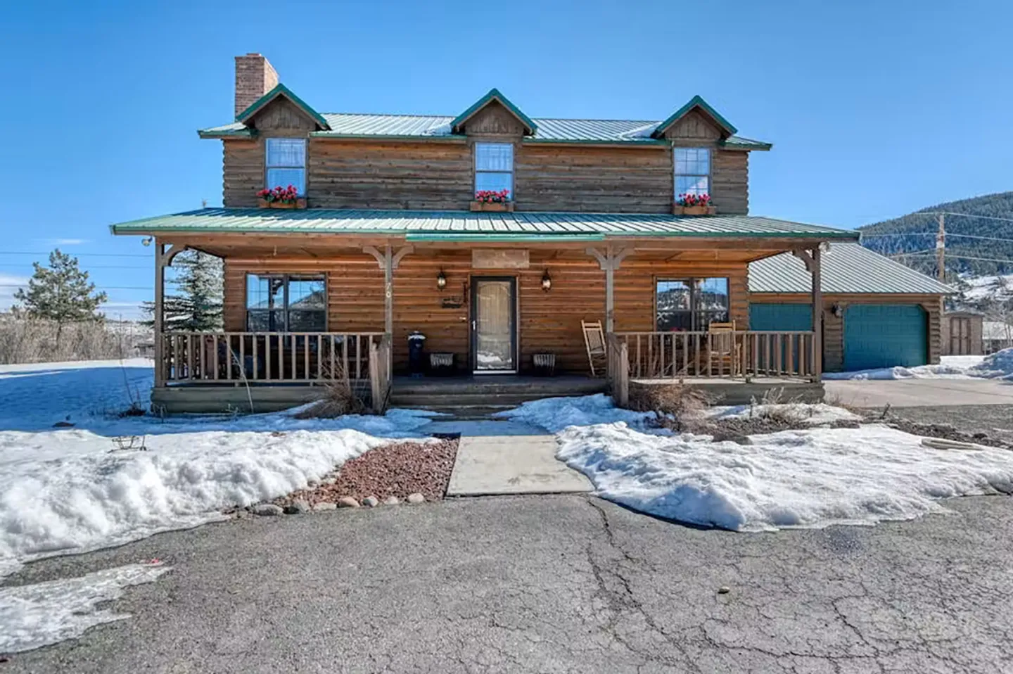 Exterior front view of a two-story log cabin with a covered front porch, green roof, and snow on the ground under a clear blue sky.