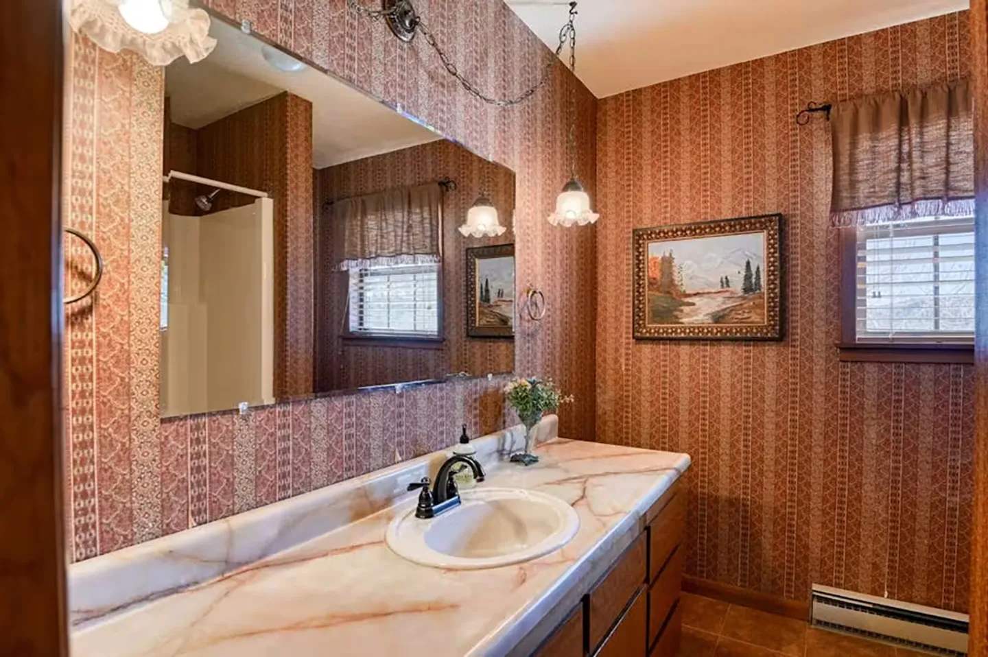 Bathroom vanity with a large mirror, patterned wallpaper, and a framed painting on the wall.
