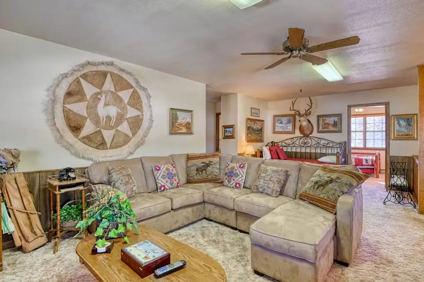 Living area with a tan sectional sofa, patterned pillows, ceiling fan, and carpet, opening to a bedroom area in the background.