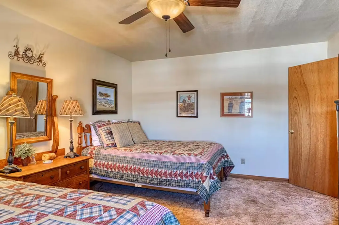 Bedroom with two beds topped with patterned quilts, a wooden dresser with a mirror, carpeted flooring, and a ceiling fan.