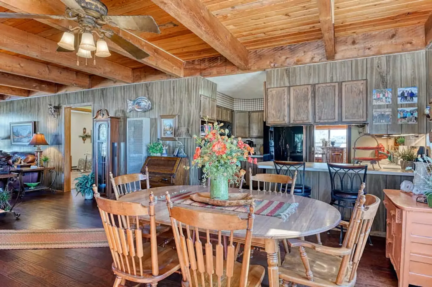 Dining area with a wooden table and chairs beneath exposed wood ceiling beams, adjacent to wood-paneled walls and a kitchen counter.