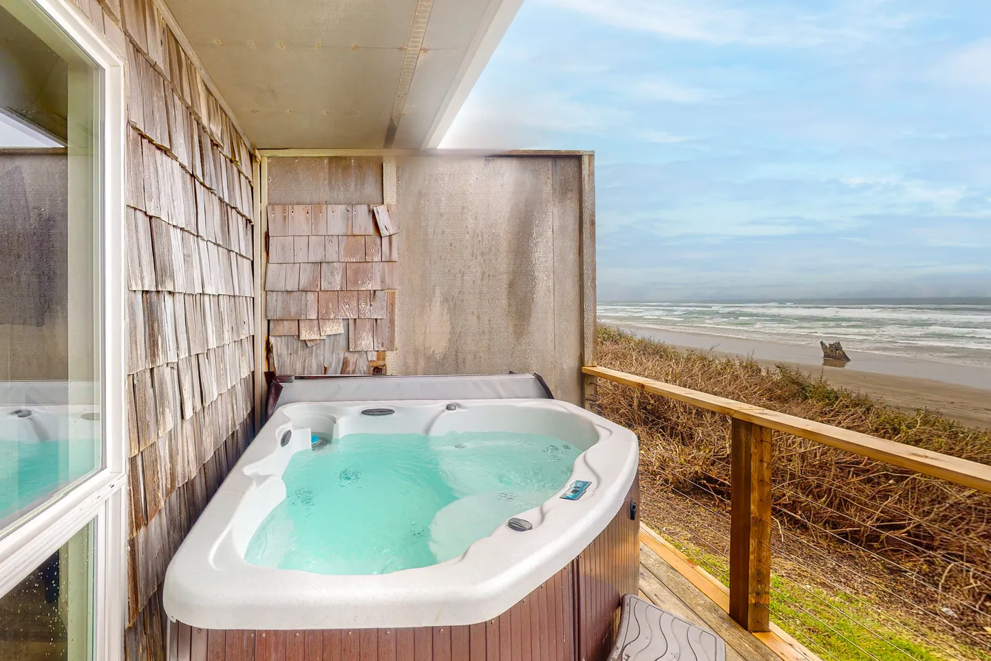 A hot tub on a wooden deck overlooking a sandy beach and ocean waves.