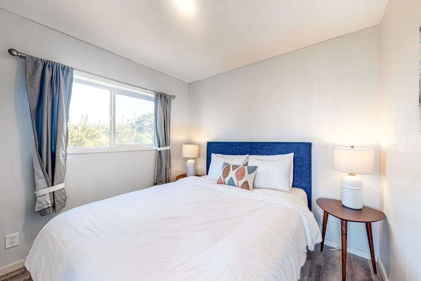 Bedroom with a bed featuring a blue upholstered headboard, white linens, a window with curtains, and bedside tables with lamps.