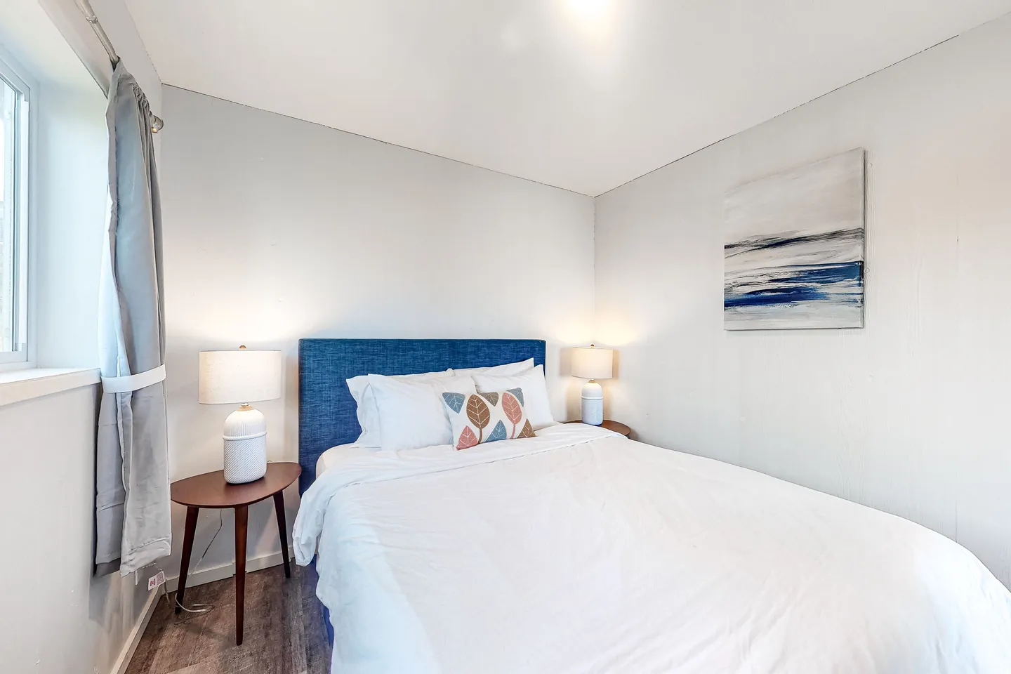 Bedroom with a blue upholstered headboard, white bedding, two bedside tables with lamps, and a window to the left.