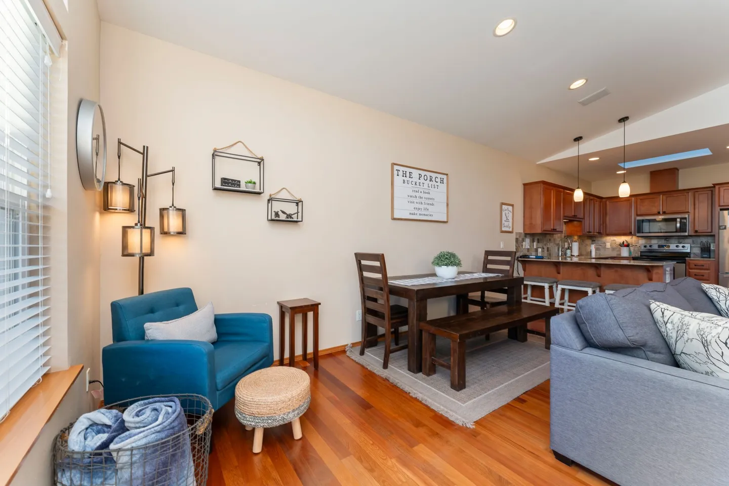 Open-concept living and dining area featuring hardwood floors, a teal armchair, a wooden dining table with a bench, and a view into the kitchen.