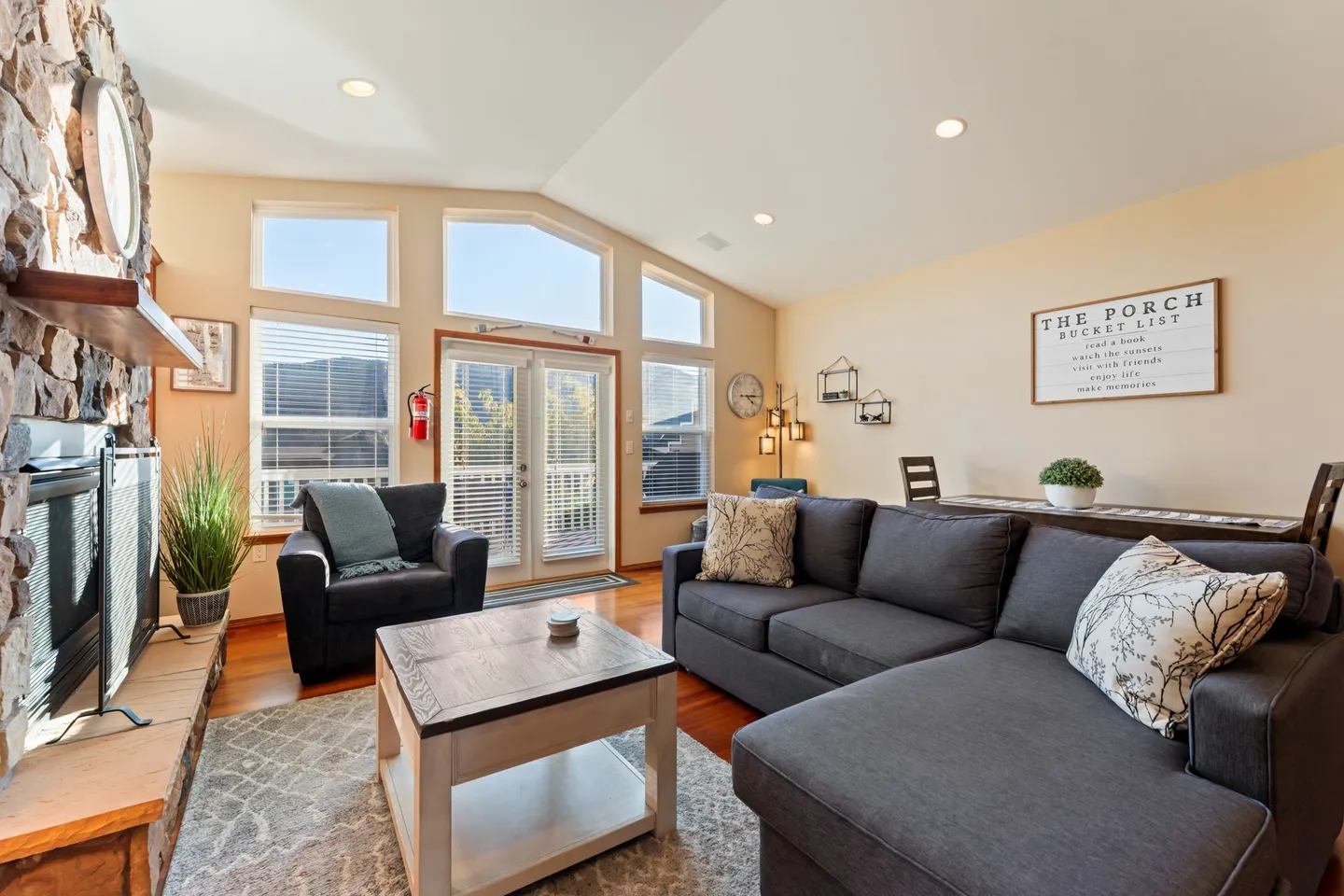 Living room featuring a dark gray sectional sofa, a coffee table, a stone fireplace, and high windows above French doors.