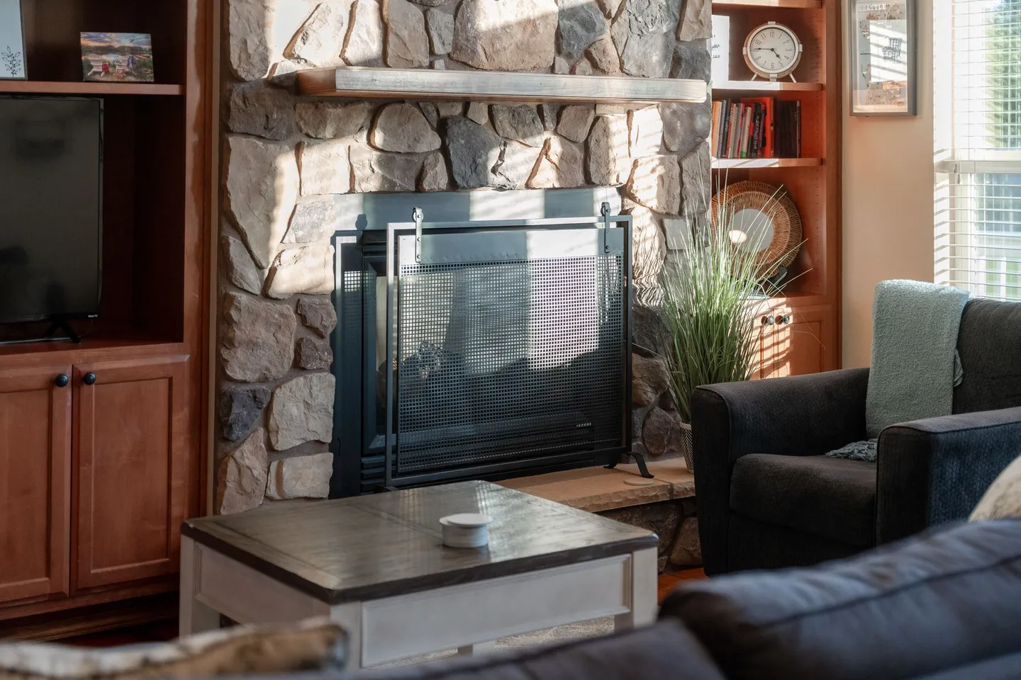 Living room featuring a stone fireplace with a wooden mantel, flanked by built-in wood shelving, a coffee table, and an armchair.