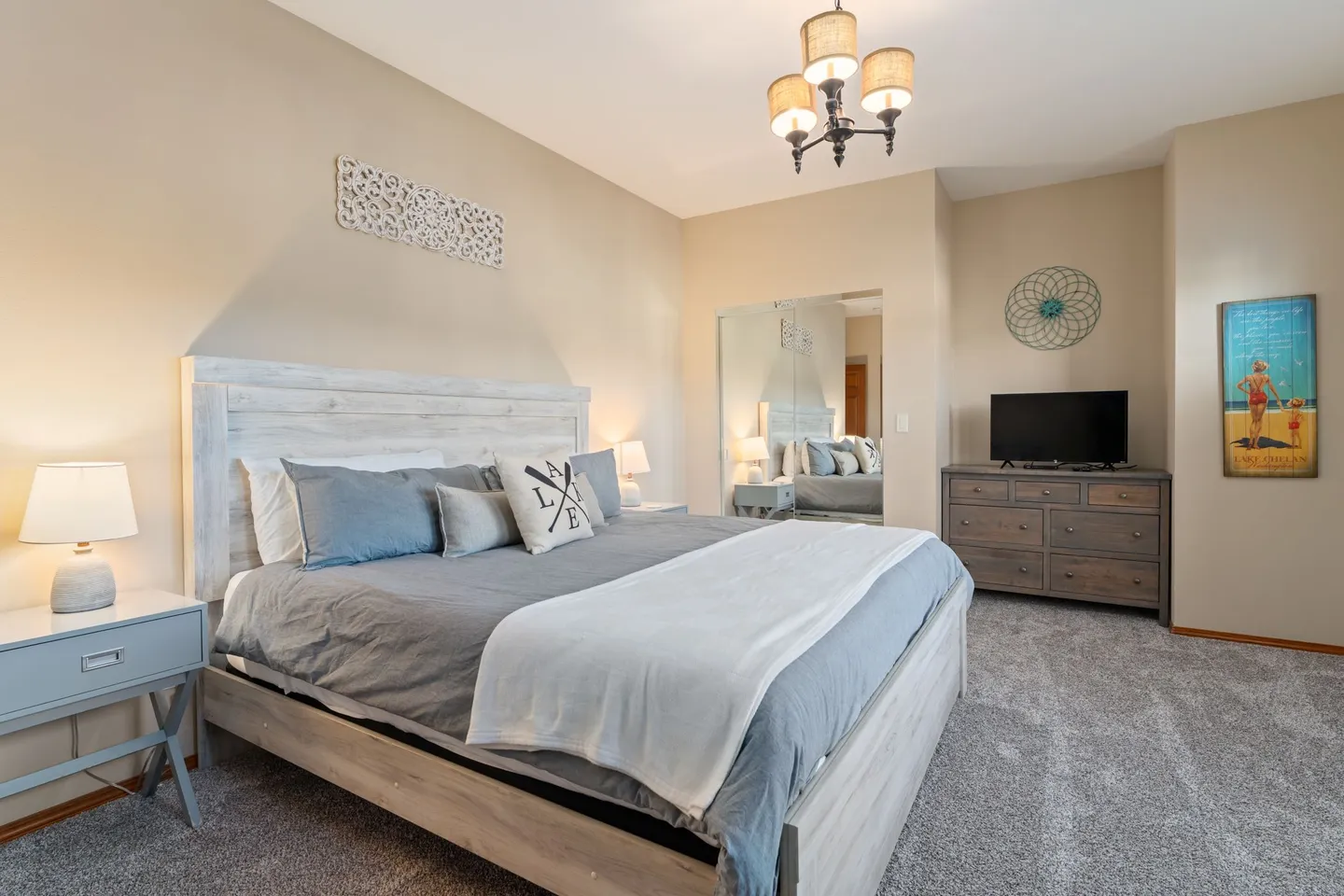 Bedroom featuring a made bed with a distressed wood headboard, bedside tables with lamps, mirrored closet doors, and a TV atop a dresser.