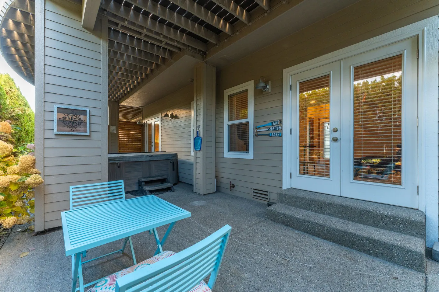 Covered concrete patio with a light blue outdoor table and chairs, adjacent to double glass doors and a hot tub under the deck.