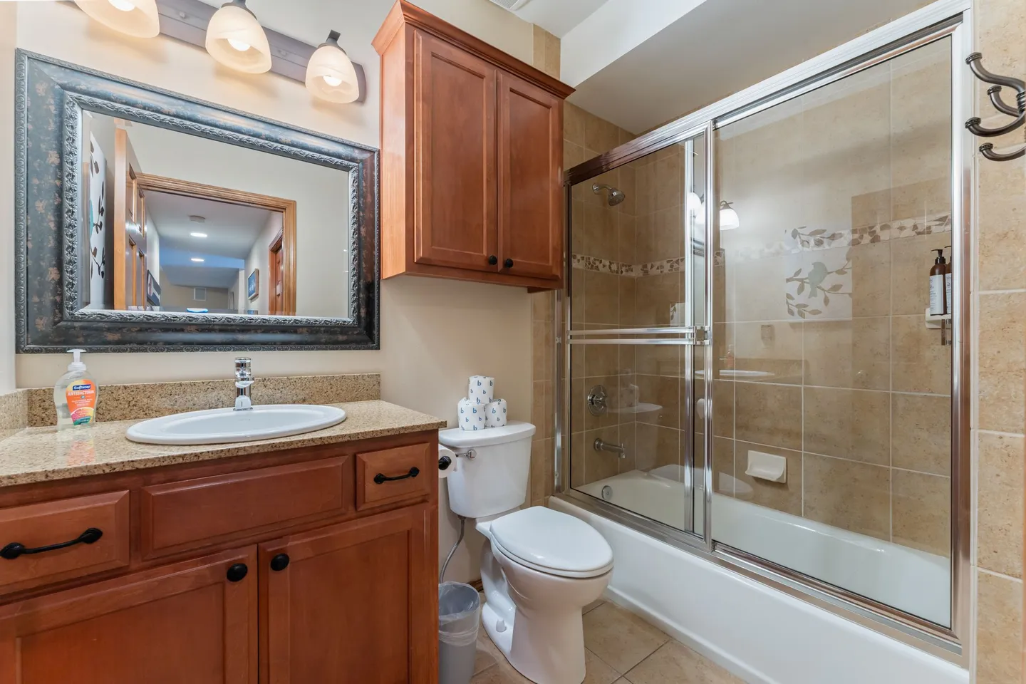 Bathroom featuring a wood vanity with an oval sink, a toilet, a wall-mounted cabinet, and a bathtub with sliding glass doors and tiled surround.