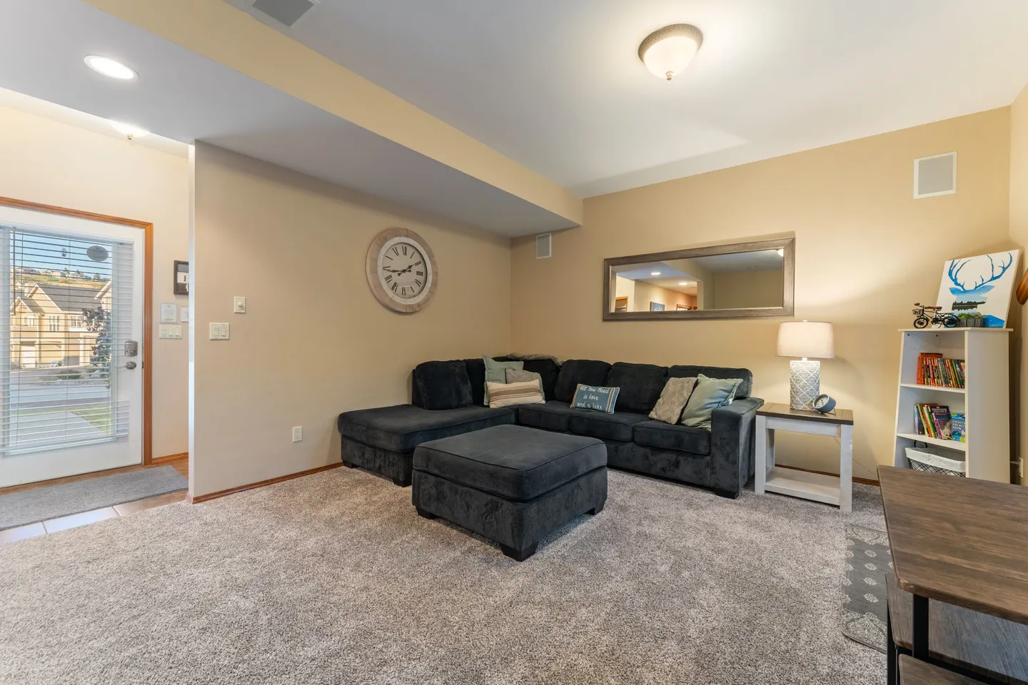 Living room with carpet flooring, a dark sectional sofa with matching ottoman, end table, and front door view.