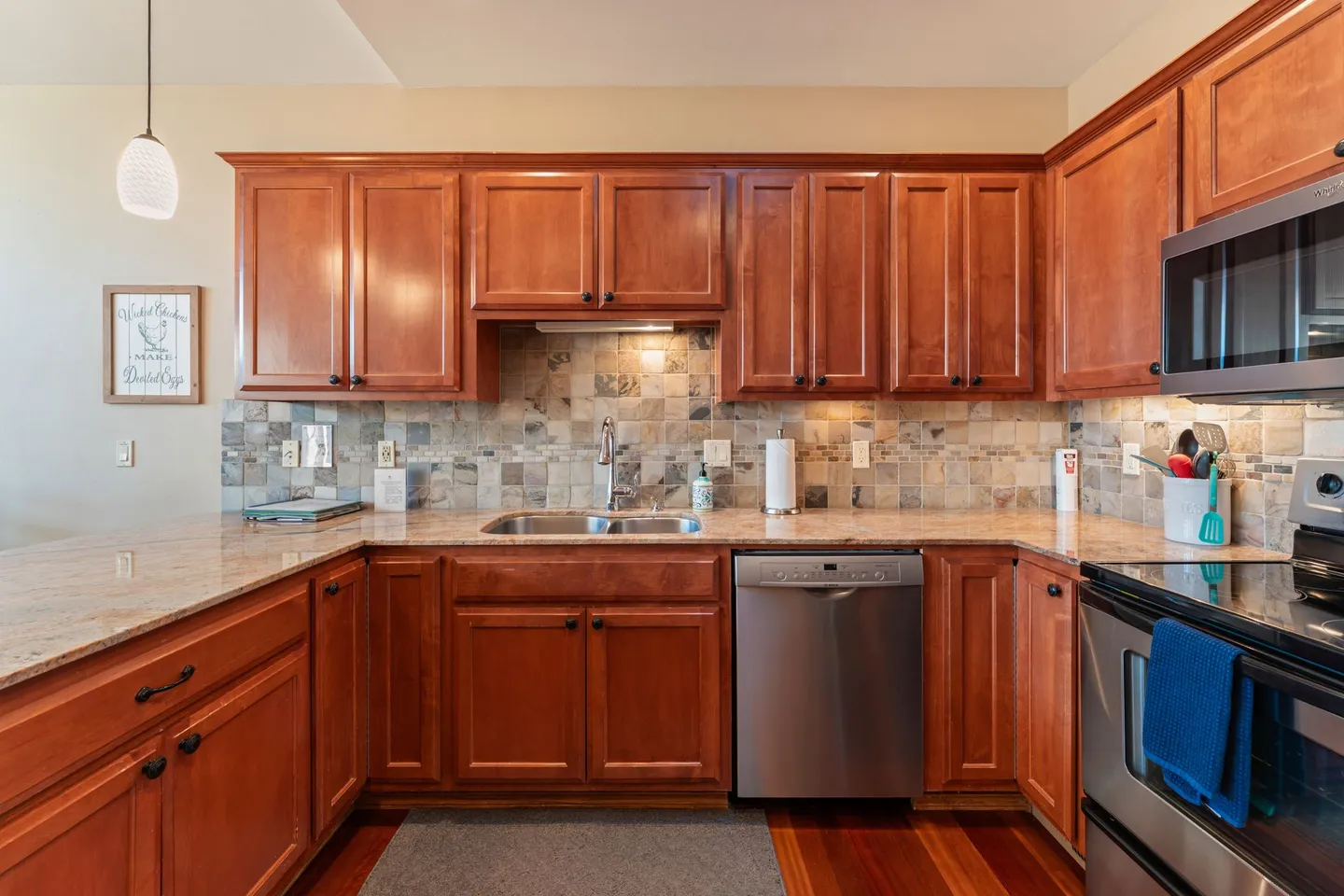 Kitchen with warm wood cabinetry, speckled countertops, stainless steel dishwasher, and tiled backsplash.